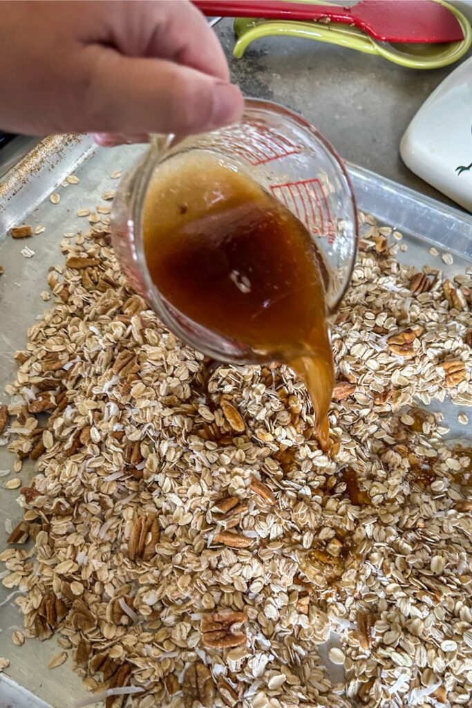 Adding maple syrup to a baking sheet of pecans and oats. 
