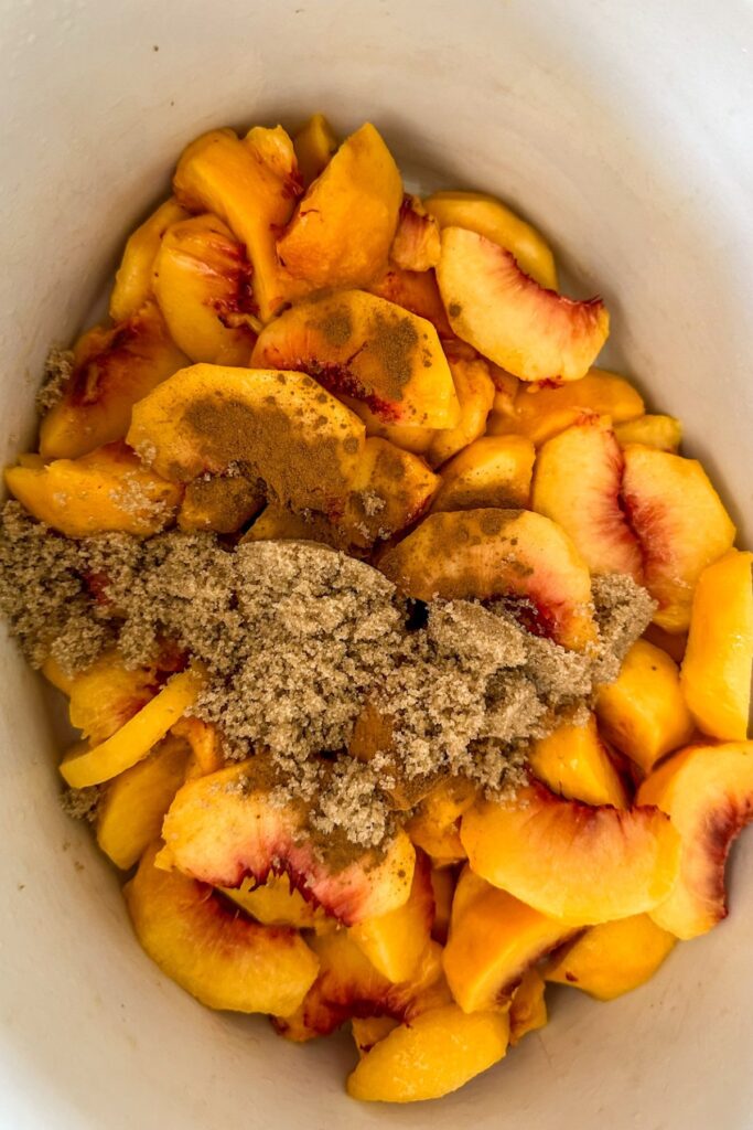Adding brown sugar and cinnamon onto sliced peaches in a Crock Pot.