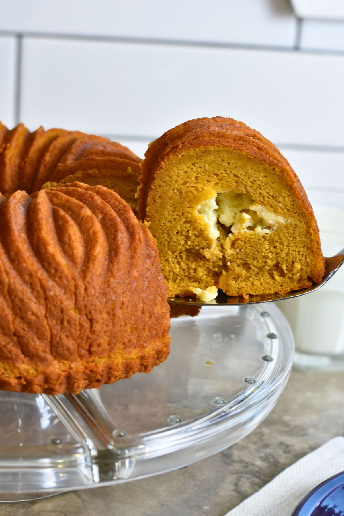 Pumpkin-cheesecake-cake-on-a-glass-cake-plate serving a slice of pumpkin cheesecake cake with a serving utensil.